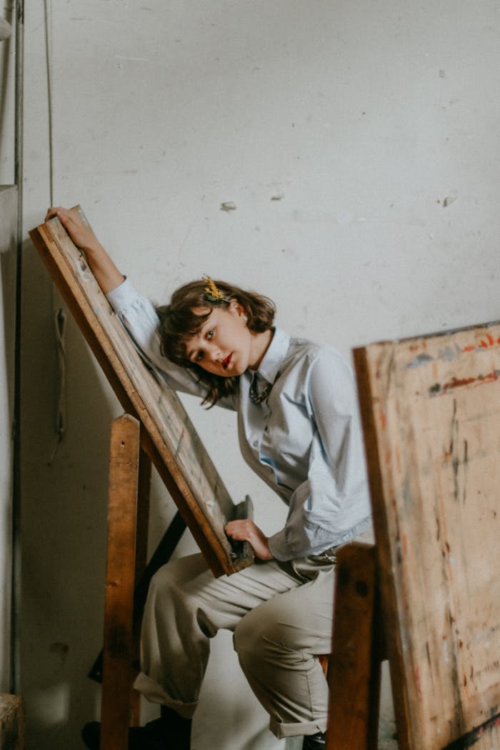 A young woman sits pensively, leaning on an easel in a cozy art studio setting.